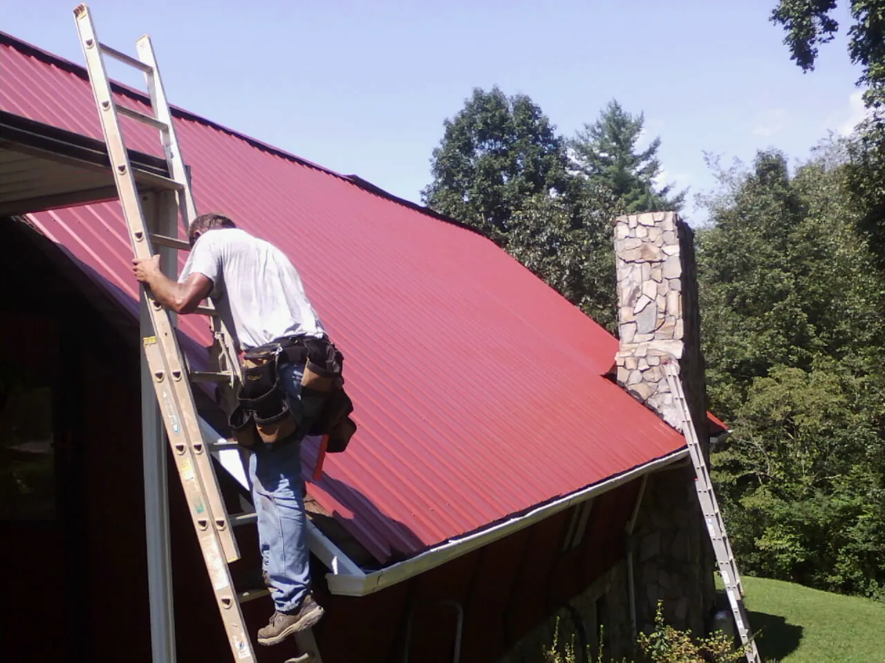 Roofer on a ladder beside a red metal roof and stone chimney — Soffit & Fascia Repair by Roofing Brothers in Parkhill